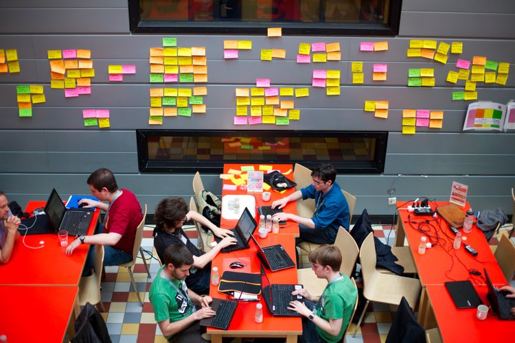 A group of white men working on a bright red table with black laptops and sticky notes.