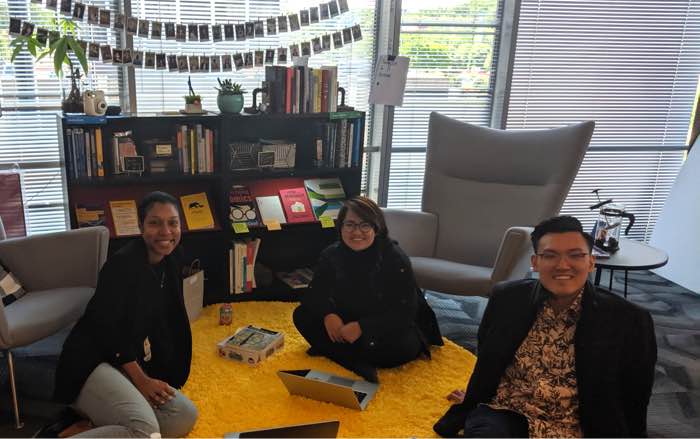 Three of my friends sitting in front of two bookshelves full of design books. There is a board game spread out on the rug we are sitting on.