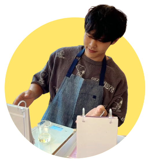 A Korean man wearing a snoopy shirt and an apron, trying to make a candle, having no clue as to what he's doing.